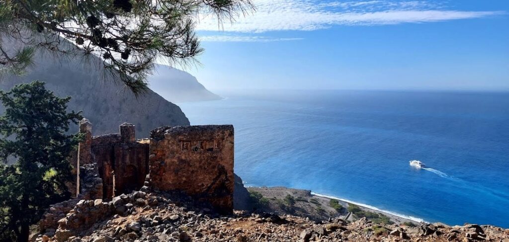 Ruins of a stone building on a rocky cliff overlooking the blue sea, with a pine branch overhead framing the view.