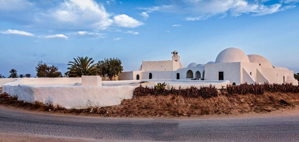 Whitewashed building complex with domed roofs and arched entryways set against a blue sky and palm trees in a desert landscape.