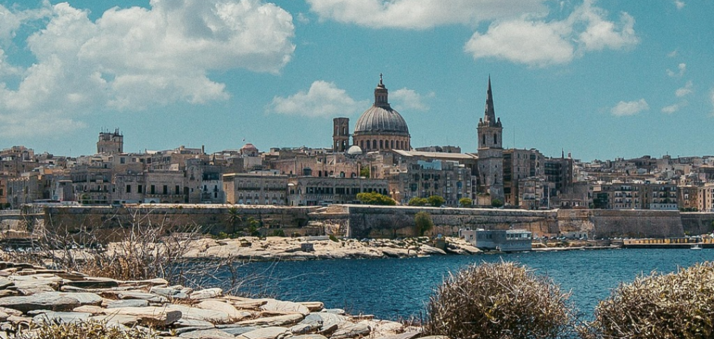 Coastal Mediterranean city skyline with a domed cathedral and church spire across the harbor, seen from a rocky foreground.