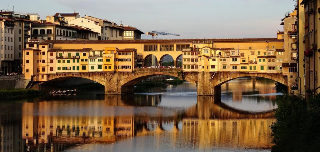 Historic stone bridge with arches crossing a calm river, lined by colorful riverside buildings at sunset.