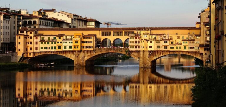 Historic stone bridge with arches crossing a calm river, lined by colorful riverside buildings at sunset.