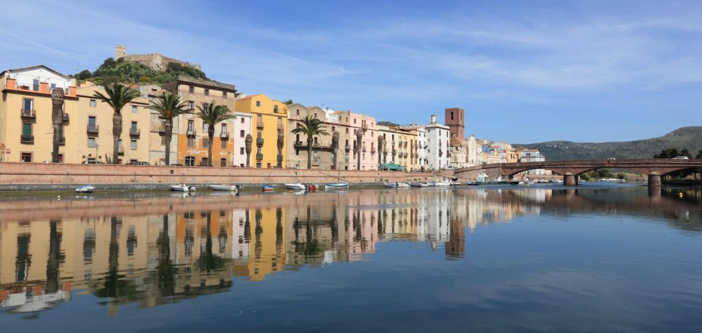 Colorful buildings along a riverfront with palm trees, a brick bridge, and distant hills under a blue sky.