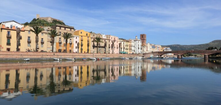 Colorful buildings along a riverfront with palm trees, a brick bridge, and distant hills under a blue sky.