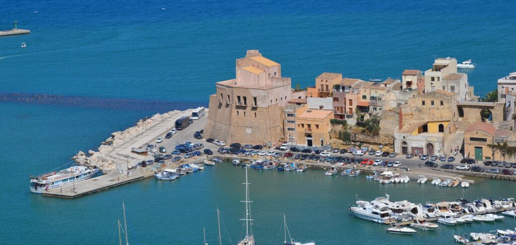 Aerial view of a coastal harbor town with stone buildings, piers, and many moored boats against blue water.
