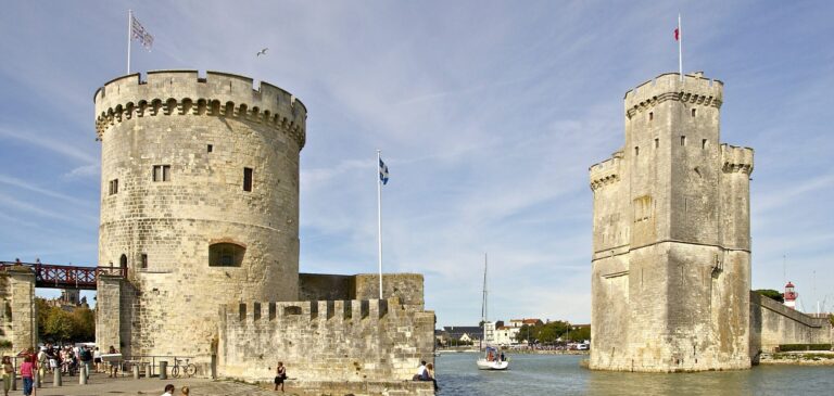Two medieval stone towers by a harbor with flags, as people stroll along the waterfront.