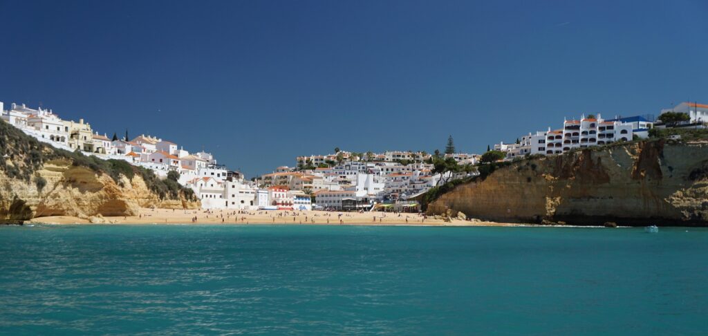 Coastal town with white buildings on cliffs overlooking a sandy beach and turquoise sea.