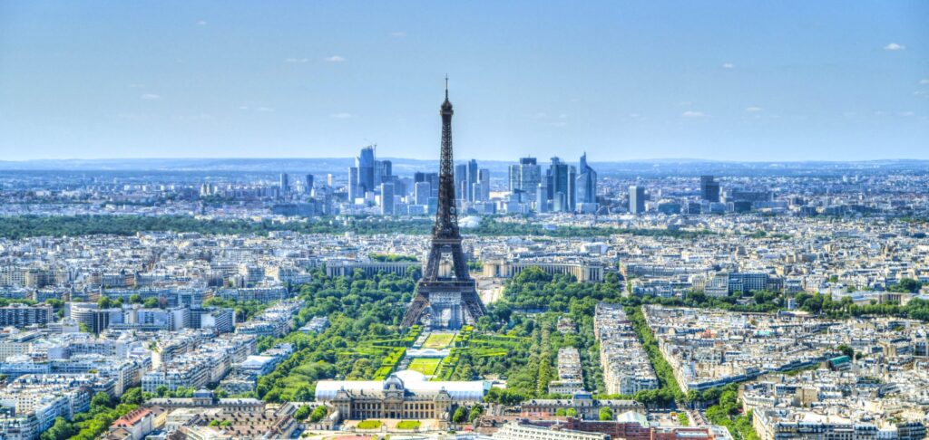 Paris cityscape with the Eiffel Tower rising above densely packed buildings and green parks under a clear blue sky.