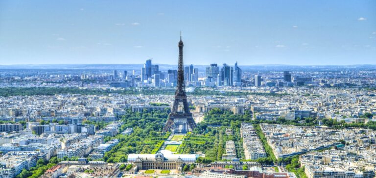 Paris cityscape with the Eiffel Tower rising above densely packed buildings and green parks under a clear blue sky.