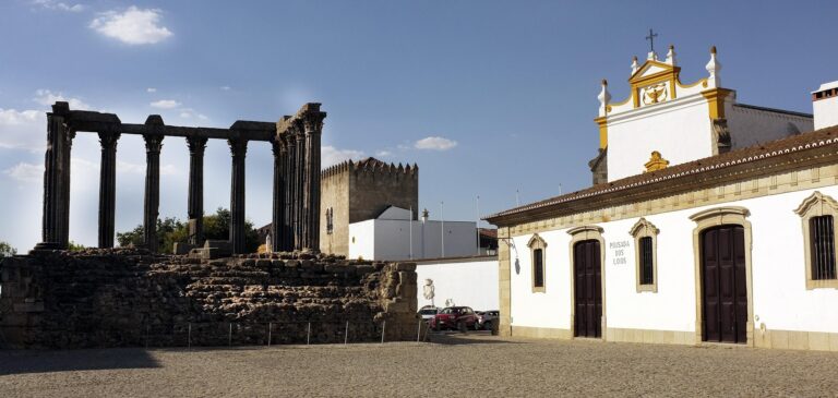 Ancient stone ruin with tall columns beside a white and yellow-trimmed building under a blue sky.