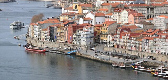 Colorful waterfront buildings with red-tiled roofs along a river, with boats docked at a stone quay.