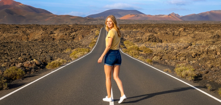 A smiling woman in denim shorts and a beige shirt stands in the middle of a long, straight road through a rocky desert landscape with mountains in the distance.