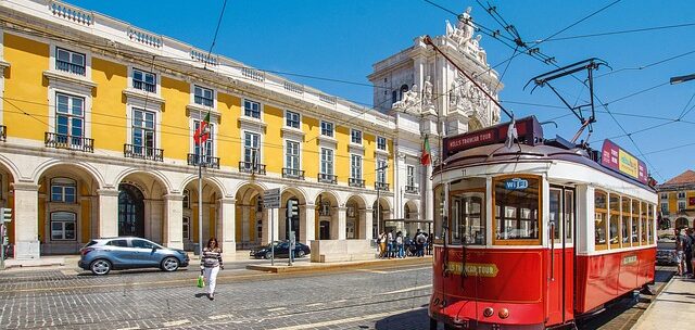Vintage red tram on a cobblestone street in a sunny city square, with a yellow arched building in the background.