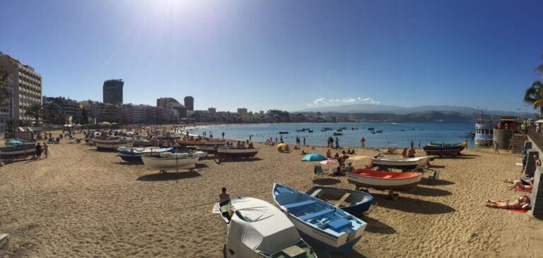 Panoramic beach scene with boats on the sand, sunbathers and swimmers in the water, and a city shoreline under a bright blue sky.