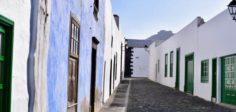 Sunny narrow street lined with white buildings; a blue wall on the left with wooden windows and a green door, and green-framed windows on the right under a clear blue sky.