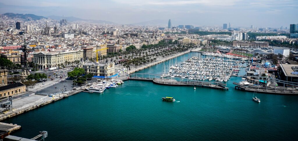 Aerial view of Port Vell harbor in Barcelona with a marina full of yachts and a palm-lined waterfront promenade against the city skyline.